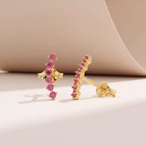 May include: A pair of gold-colored stud earrings. Each earring features a line of small, round, pink gemstones. One earring has a unique, geometric design at the base, while the other has a straight post.