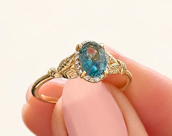 Könnte beinhalten: Ein goldener Ring mit einem oval geschliffenen blau-grünen Edelstein in der Mitte, umgeben von einem Kranz aus kleinen weißen Steinen. Die Ringschiene ist auf beiden Seiten der Fassung mit detaillierten Blattgravuren versehen.