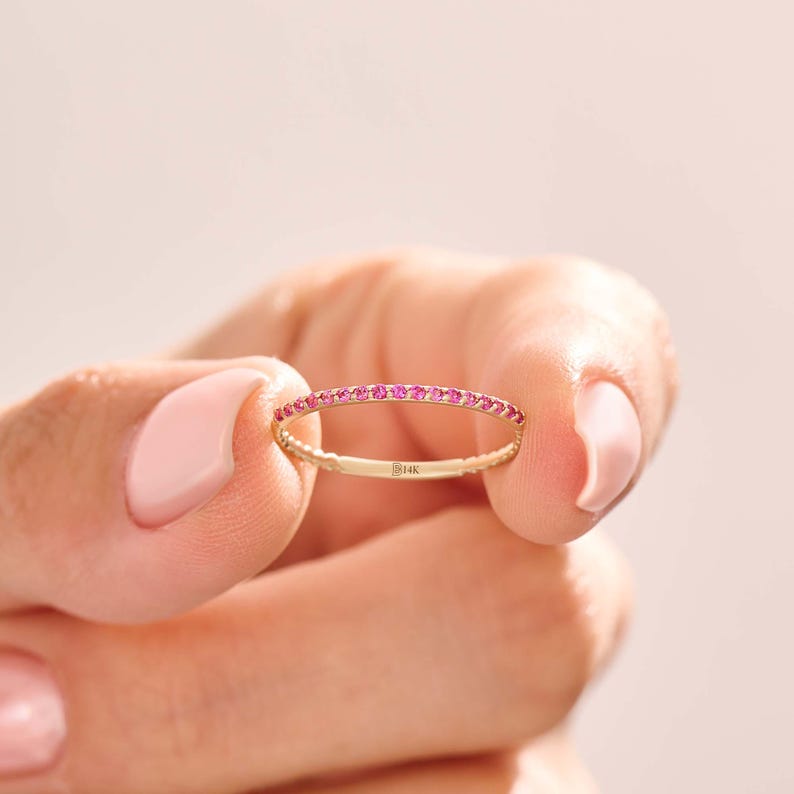 Puede incluir: Un delicado anillo de oro con una banda de peque&ntilde;as y vibrantes piedras preciosas rosas. El anillo se sostiene entre dos dedos sobre un fondo rosa suave. El anillo parece tener aproximadamente 0,25 cm de ancho.