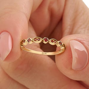 May include: A gold ring featuring a line of small, red gemstones. The ring's design includes a repeating pattern of gemstones and decorative details. The ring is held between two fingers. The ring is marked with 'B14K'.