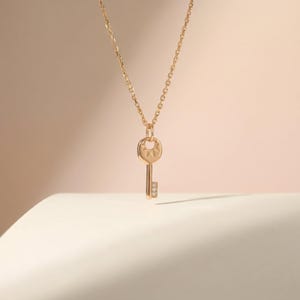 May include: A gold-coloured key pendant necklace. The key charm features a sunburst design on the head and three small clear stones on the stem. The necklace chain is delicate and gold-toned. The background is a soft, neutral colour.