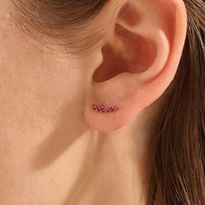 May include: A gold ear climber earring featuring a curved row of five small, round, vibrant pink gemstones. The earring is positioned along the lower curve of the earlobe, showcasing a delicate and minimalist design with a polished metal finish.