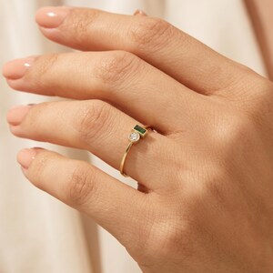 May include: A thin gold ring featuring two bezel-set stones: a small round clear white gemstone and a rectangular baguette-cut green gemstone positioned side-by-side on a delicate polished gold band that encircles the finger.