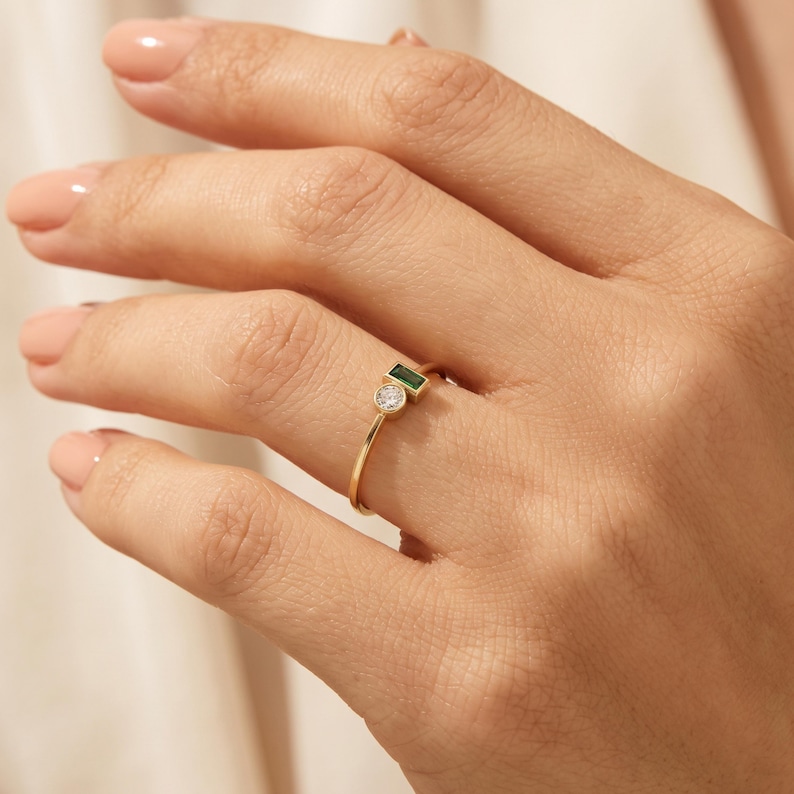 May include: A thin gold ring featuring two bezel-set stones: a small round clear white gemstone and a rectangular baguette-cut green gemstone positioned side-by-side on a delicate polished gold band that encircles the finger.