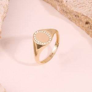 May include: A gold signet ring with a smooth oval top surrounded by a row of small diamonds. The ring is marked "B 14K" on the inside band.