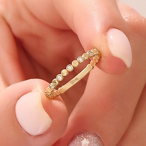 May include: A gold ring with a series of small, round, clear stones set in a repeating pattern. The ring band is smooth and bears the inscription "B14K". The ring is held between two fingers.