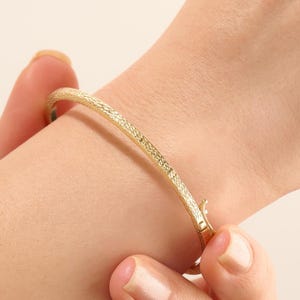 Könnte beinhalten: Ein goldener Armreif mit einer texturierten, kreuzschraffierten Oberfläche. Der Armreif ist mit einem Scharnierverschluss ausgestattet, der die beiden Enden miteinander verbindet und so ein durchgehendes, rundes Band bildet.