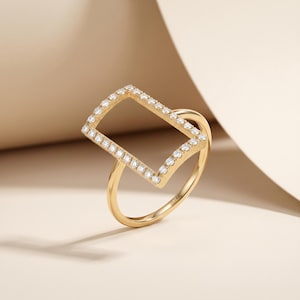 May include: A gold ring with a rectangular design encrusted with small, clear gemstones. The ring has a delicate band and a geometric shape, showcasing a modern and elegant style. The ring is set against a neutral background.