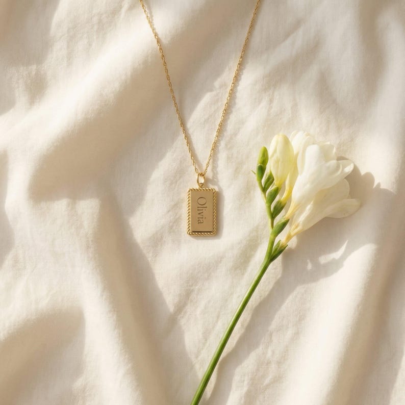 May include: A gold-toned necklace featuring a rectangular pendant with a rope-style border. The pendant is engraved with the name Olivia in a serif font. The necklace hangs from a delicate gold-toned chain, accompanied by a white freesia flower.