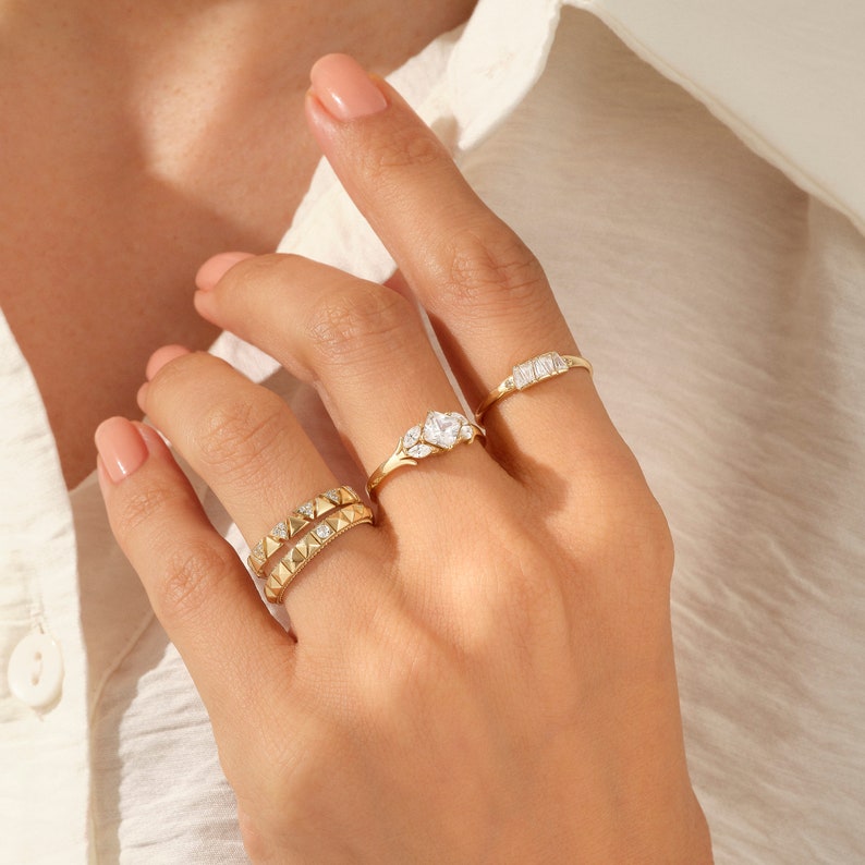 May include: Three gold rings of varying designs are shown on a hand. One ring has a row of pyramid-shaped studs, another features a heart-shaped stone, and the third has a rectangular stone design. The rings are set against a neutral background.