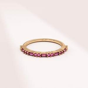 Op de afbeelding: Een gouden bandring met een rij rechthoekige, roze edelstenen. De ring heeft een delicaat ontwerp, met de edelstenen dicht bij elkaar rond de band. De ring wordt getoond tegen een witte achtergrond.