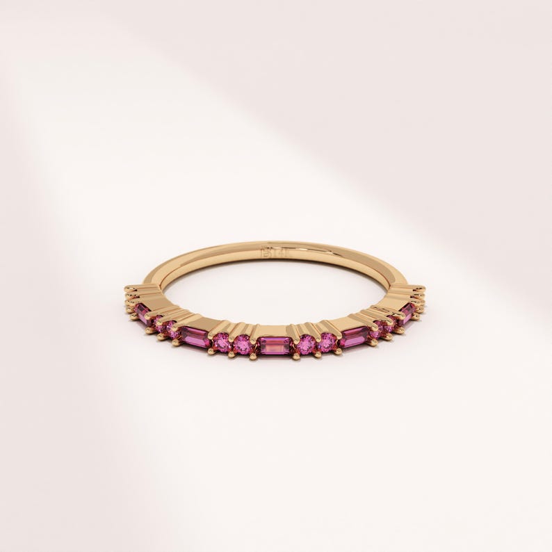 Op de afbeelding: Een gouden bandring met een rij rechthoekige, roze edelstenen. De ring heeft een delicaat ontwerp, met de edelstenen dicht bij elkaar rond de band. De ring wordt getoond tegen een witte achtergrond.