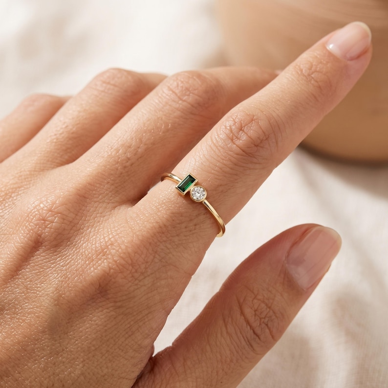 May include: A gold ring featuring a small rectangular emerald-cut green gemstone and a single round brilliant-cut clear gemstone set side-by-side on a thin, polished gold band. The stones are positioned horizontally along the finger.