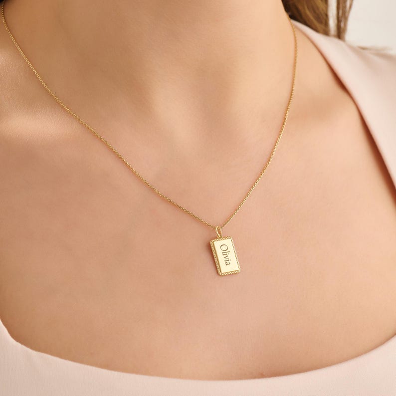 May include: A gold-toned necklace featuring a delicate chain and a rectangular pendant with a rope-style border. The pendant is engraved with the name Olivia in a clear, centred font, creating a simple and refined piece of jewellery.