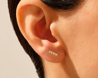 May include: A close-up of a person's ear with a gold bar earring featuring five small round beads.
