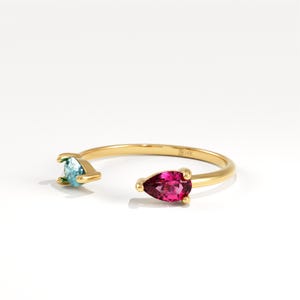 May include: Open-style gold ring with a pear-shaped, bright fuchsia gemstone and a light blue gemstone. The ring has a slender band and is set against a white backdrop. The ring is marked with "14K".