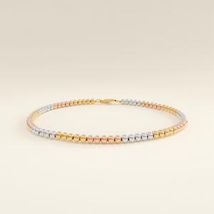 Könnte beinhalten: Eine zarte Halskette mit einem Farbverlauf aus goldfarbenen, roségoldenen und silberfarbenen Perlen. Die Perlen sind gleichmäßig angeordnet und durch einen goldenen Verschluss verbunden. Der Schmuck wird vor einem neutralen Hintergrund präsentiert.