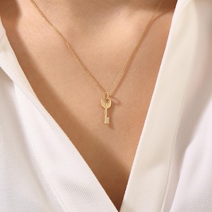 May include: A gold-colored key pendant necklace. The key charm has a detailed design and a small gemstone at the bottom. The necklace is worn against a white shirt.