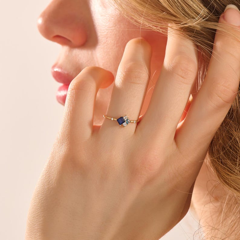 May include: A delicate gold ring featuring a square, dark blue gemstone at its center, flanked by smaller, lighter blue and clear stones. The ring's thin band is adorned with tiny accent stones, creating a refined and elegant look. The ring is on a person's finger.