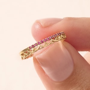 May include: A gold ring featuring a row of small, vibrant pink gemstones set along the top edge. The band is crafted with an intricate, openwork scalloped design that runs beneath the gemstone setting, creating a delicate and detailed appearance.
