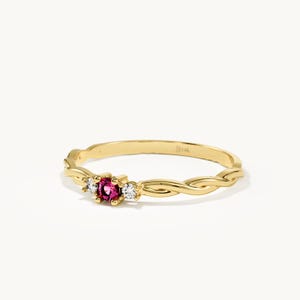 May include: A 14K yellow gold ring featuring a central round-cut pink gemstone flanked by two smaller clear round-cut stones. The band has a twisted, rope-like design and is stamped with 14K on the inner surface of the metal band.