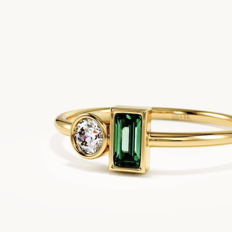 May include: A 14k gold ring featuring a round brilliant-cut clear stone in a bezel setting next to a rectangular baguette-cut green stone in a bezel setting. The band is thin and polished gold, with the 14k hallmark clearly visible on the inner surface.