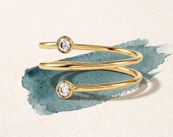 May include: A gold bypass ring featuring a spiral design with two round, clear, faceted stones set in bezel settings at each end of the band. The ring has a polished, smooth finish and a continuous, open-ended coiled structure.