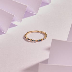 Könnte beinhalten: Ein goldener Ring mit einem Band aus kleinen, dunkelblauen Edelsteinen. Der Ring ist vor einem weichen, hellen Hintergrund mit einem geometrischen Papierdesign platziert.