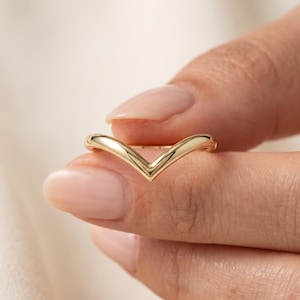 May include: A polished gold ring featuring a V-shaped chevron design. The band has a smooth, rounded surface and a continuous metallic finish, creating a simple and clean aesthetic. The ring is held between two fingers, showcasing its shape.