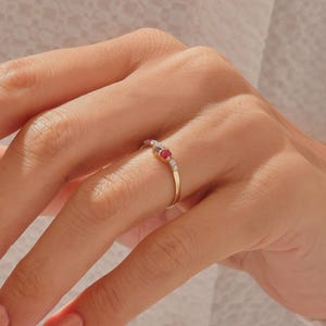 May include: A thin gold band ring featuring a central cluster of small, round-cut gemstones. The setting includes a vibrant red stone flanked by smaller, clear white stones on either side, creating a delicate and refined piece of jewelry.