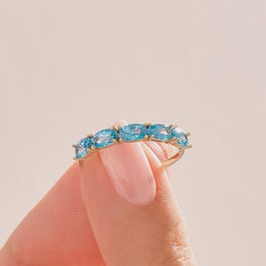 May include: A gold ring featuring five oval-cut, light blue gemstones. The ring is held up against a soft pink background, showcasing the ring's design and color. The gemstones are evenly spaced and set in a gold band.