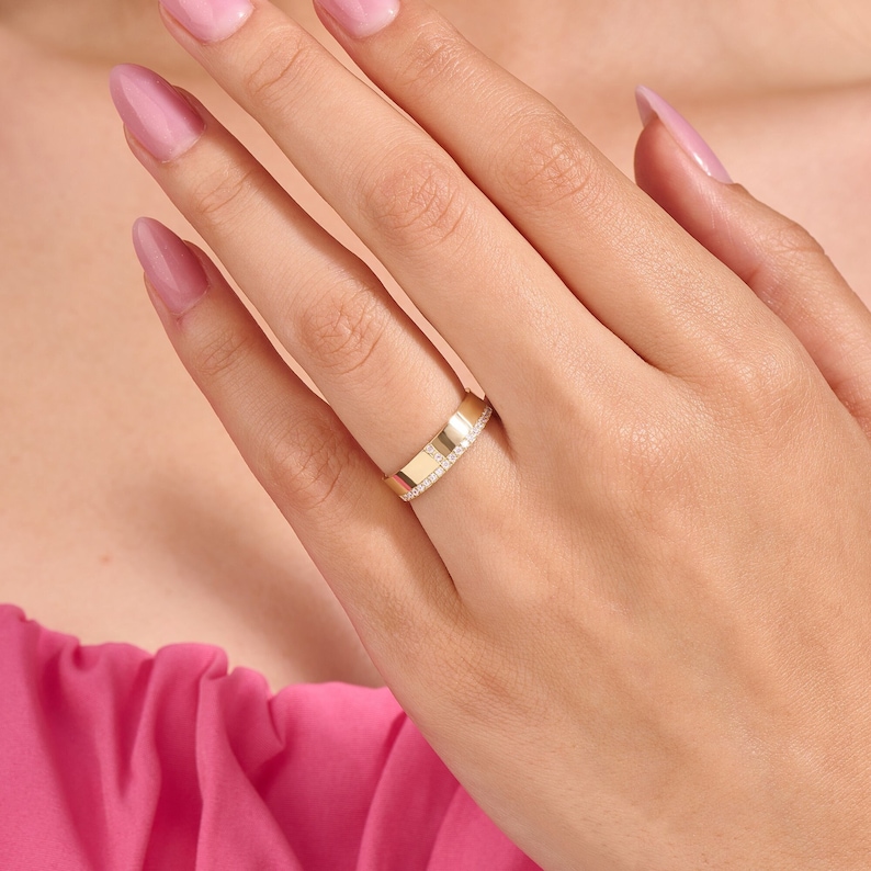 May include: A gold ring with a flat, rectangular design. One side of the ring is adorned with small, sparkling gemstones. The ring is worn on a finger, showcasing its elegant and modern style. The ring's polished surface reflects light, highlighting its luxurious appeal.