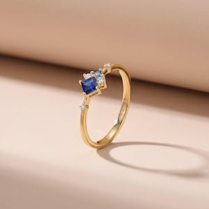 May include: A gold ring featuring a square, dark blue gemstone, flanked by smaller blue and clear stones. The ring has a delicate band and is set against a neutral background. The ring is a piece of jewelry.