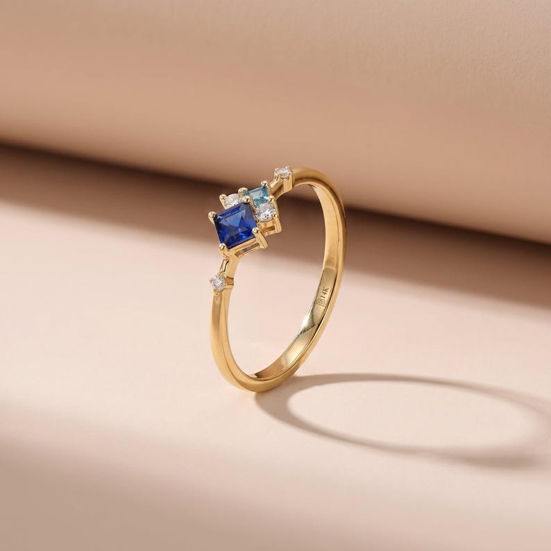 May include: A gold ring featuring a square, dark blue gemstone, flanked by smaller blue and clear stones. The ring has a delicate band and is set against a neutral background. The ring is a piece of jewelry.