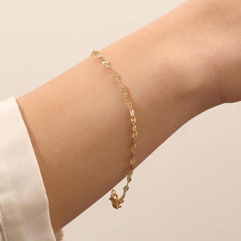 May include: A delicate gold chain bracelet featuring a series of small, flat, circular links with a textured, sunburst-like pattern. The bracelet is secured with a small lobster claw clasp, creating a refined and subtle appearance on the wrist.