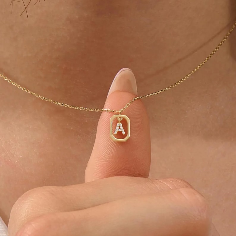 May include: A fine gold necklace with a pendant displaying the letter "A" within a rectangular setting. The pendant is embellished with tiny, glittering stones. The necklace is shown on a person's finger.
