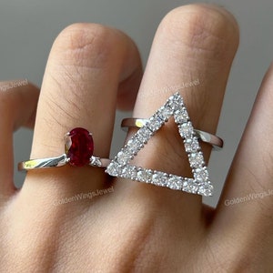 May include: Two silver rings on a hand. One ring features a red oval gemstone. The other ring is a triangle shape with small clear gemstones.