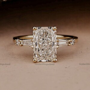 Puede incluir: Anillo de compromiso de oro con un diamante de talla radiante. El anillo presenta una gran piedra central rectangular y diamantes baguette más pequeños a cada lado. El diseño es clásico y elegante.