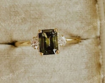 May include: A gold ring with a green emerald-cut gemstone and two smaller diamonds on either side. The ring is displayed in a white velvet ring box.