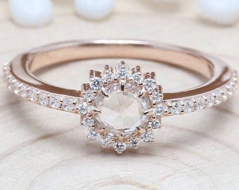 May include: A rose gold engagement ring with a round, bezel-set diamond center stone. The center stone is surrounded by a halo of smaller diamonds. The band is also set with smaller diamonds.