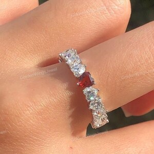 Könnte beinhalten: Ein silberner Ring mit einer Reihe von klaren Zirkonia-Steinen und einem einzigen roten herzförmigen Stein in der Mitte.