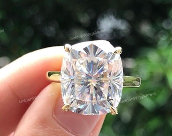 May include: A gold ring featuring a large, cushion-cut diamond. The diamond is clear and faceted, reflecting light. The ring has a simple, elegant design with a gold band and prongs holding the diamond.