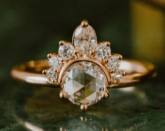 May include: Rose gold ring featuring a large, faceted, clear gemstone at the center, surrounded by smaller, clear gemstones in a crown-like design. The ring is set against a reflective, green surface. The text "Golden Wings Jewel" is visible.