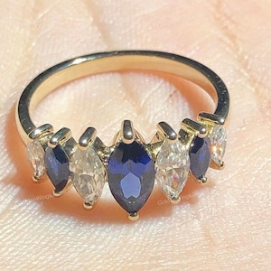 May include: A gold ring featuring a series of marquise-cut gemstones. Alternating between deep blue and clear stones, the ring has a delicate, elegant design. The gemstones are set in a row along the band, creating a sparkling effect.