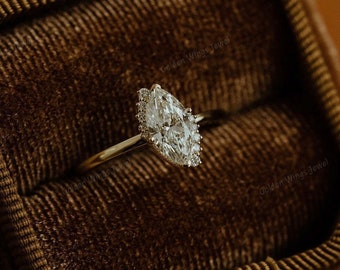 Può includere: Un anello di fidanzamento con diamante a forma di pera con una montatura a halo. L'anello è incastonato su una fascia d'oro ed è esposto in una scatola per anelli in velluto marrone.