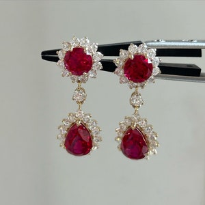 May include: A pair of dangle earrings with red ruby gemstones and a halo of clear diamonds. The earrings have a pear-shaped ruby gemstone on the bottom.