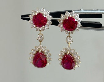 Puede incluir: Un par de pendientes colgantes con piedras preciosas de rubí rojo y un halo de diamantes transparentes. Los pendientes tienen una piedra preciosa de rubí en forma de pera en la parte inferior.