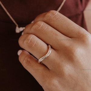 Puede incluir: Un anillo de oro curvado con piedras preciosas rectangulares. El anillo se lleva en un dedo, con un collar visible en el fondo. El diseño del anillo es elegante y moderno, con un enfoque en las piedras preciosas.