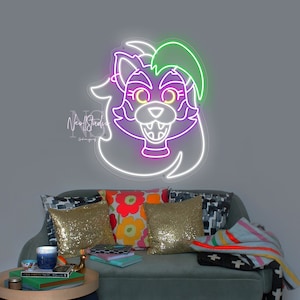 Może przedstawiać: Neonowy znak przedstawiający futrzaste zwierzę z zielonymi, fioletowymi i białymi akcentami. Znak jest zamontowany na szarej ścianie nad zieloną kanapą z kolorowymi poduszkami i kocem. Mały czarny stolik z książkami i kubkiem stoi przed kanapą.