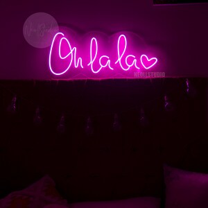 May include: A bright pink neon sign mounted on a wall, displaying the cursive text Oh la la followed by a heart symbol. The sign emits a vibrant glow, illuminating the dark space below where bedding and a string of small light bulbs are visible.